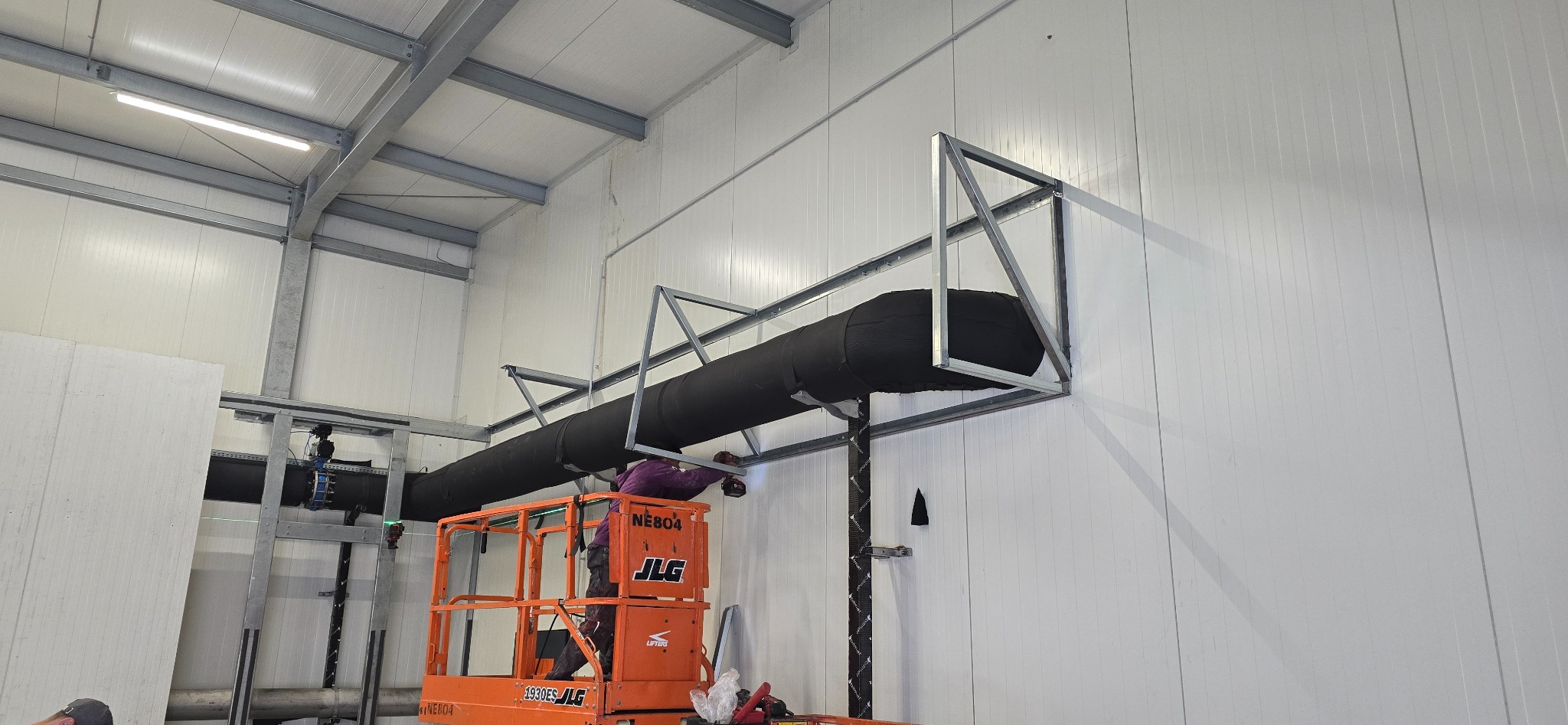 Pracownik z Lublina na podnośniku montuje metalową konstrukcję pod rurę wentylacyjną wewnątrz hali z płyt warstwowych.