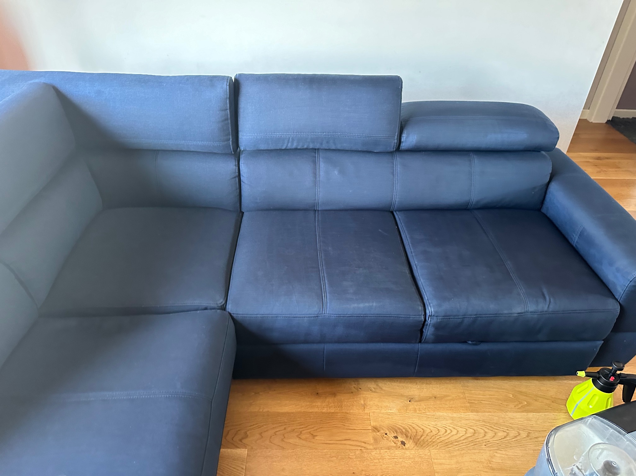 Częściowo wyczyszczona, modułowa, narożna sofa w odcieniach granatu, z widoczną różnicą w kolorze po praniu na lewym module, na podłodze leży spryskiwacz i odkurzacz piorący.
