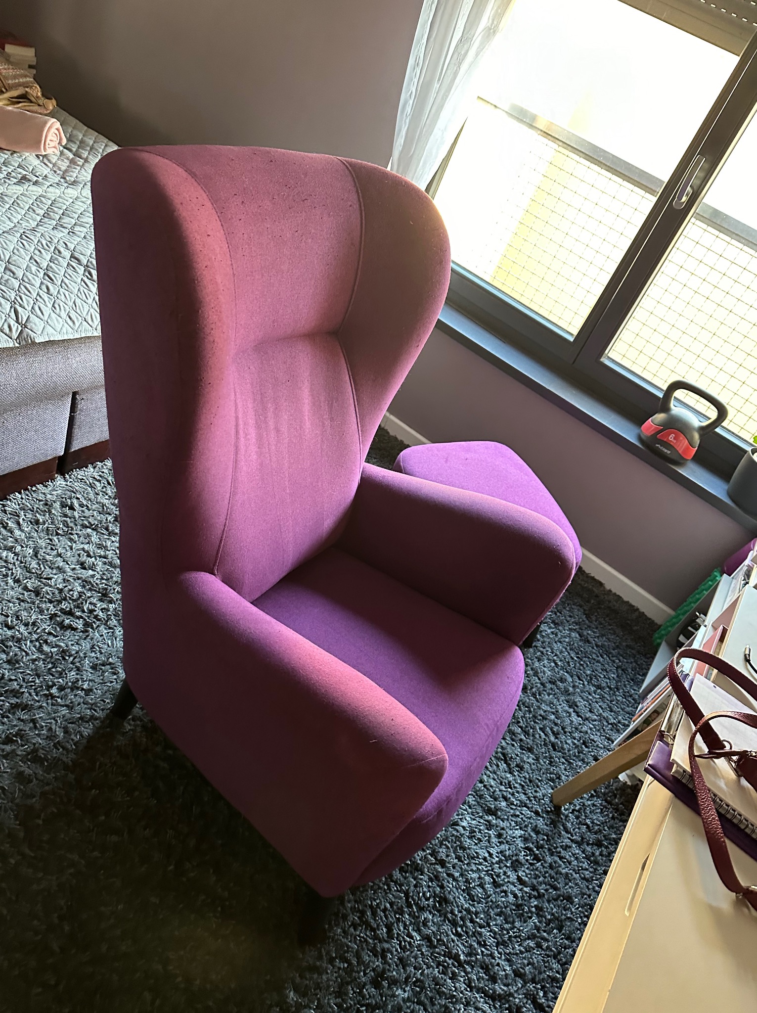 Fioletowy fotel uszak na tle szarego dywanu, obok okna z widoczną wagą kettlebell i fragmentem biurka z torbą.