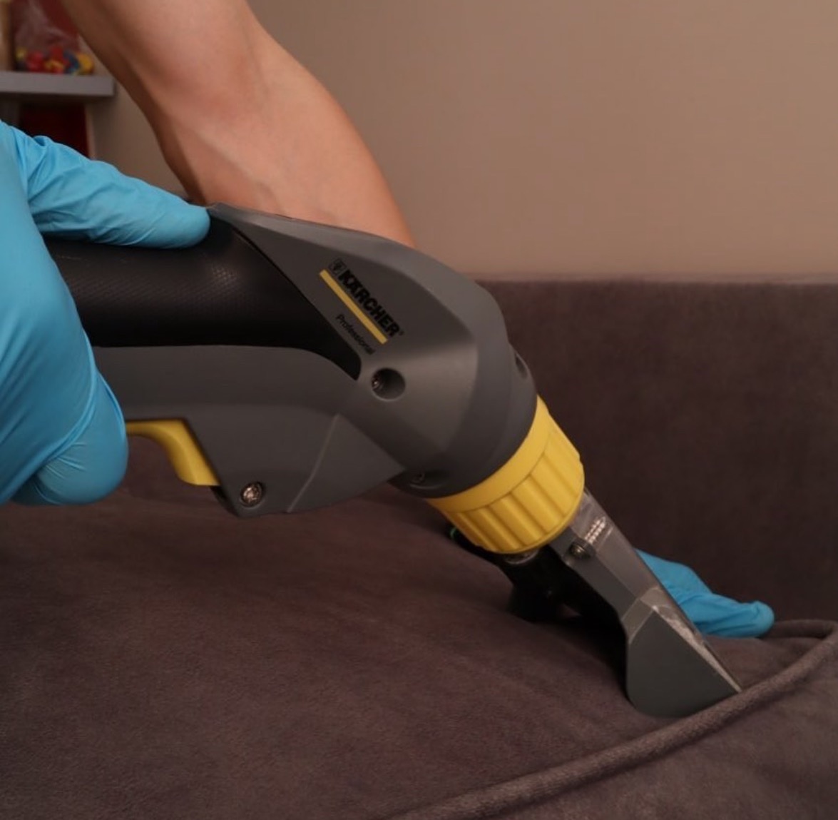 Profesjonalne urządzenie Kärcher w trakcie czyszczenia szarej sofy, widoczne błękitne rękawice ochronne.