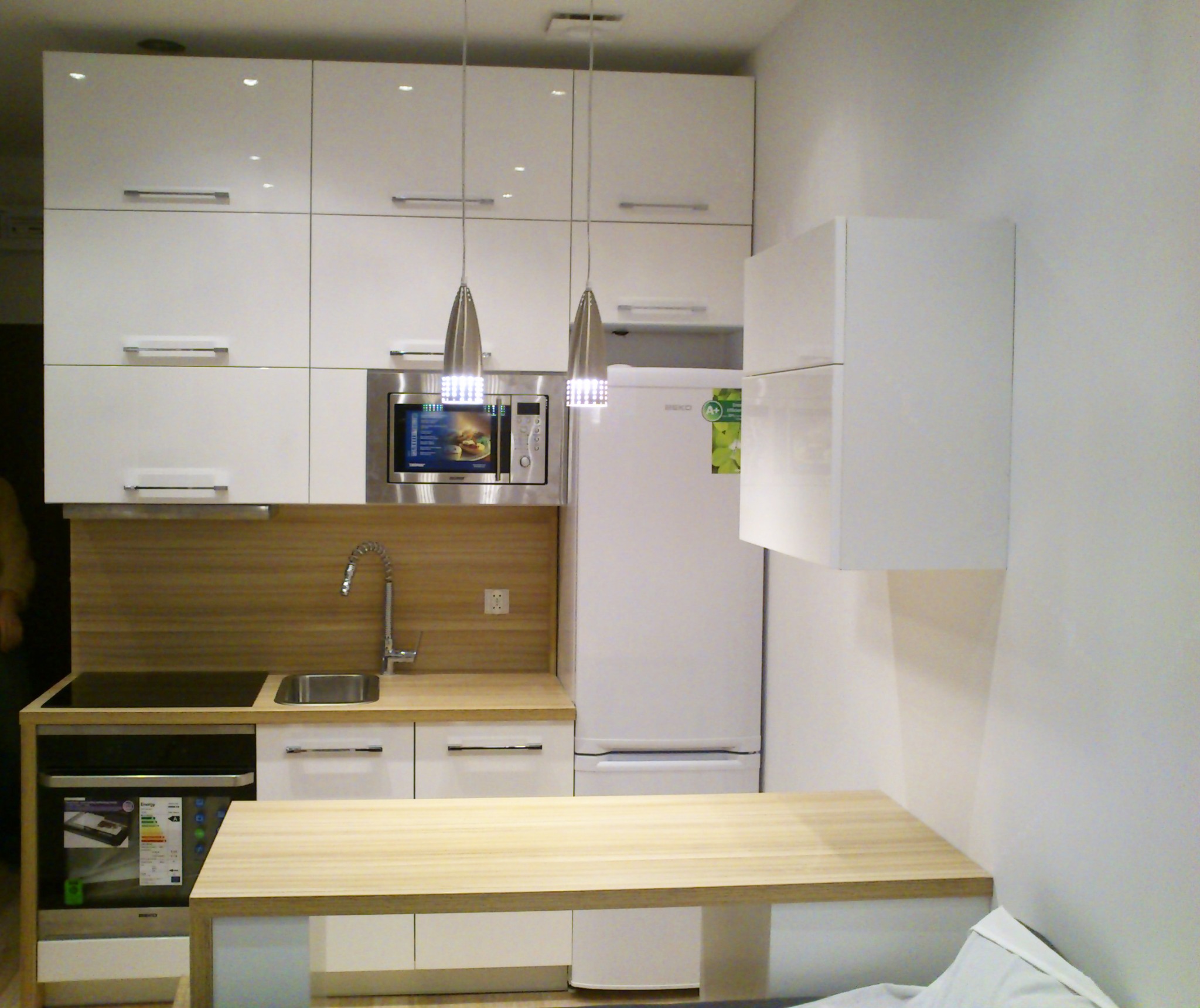 Nowoczesna, mała kuchnia z białymi, lakierowanymi szafkami, drewnianym blatem i srebrnymi lampami wiszącymi, widok na kuchenkę, zlewozmywak, mikrofalówkę i lodówkę.