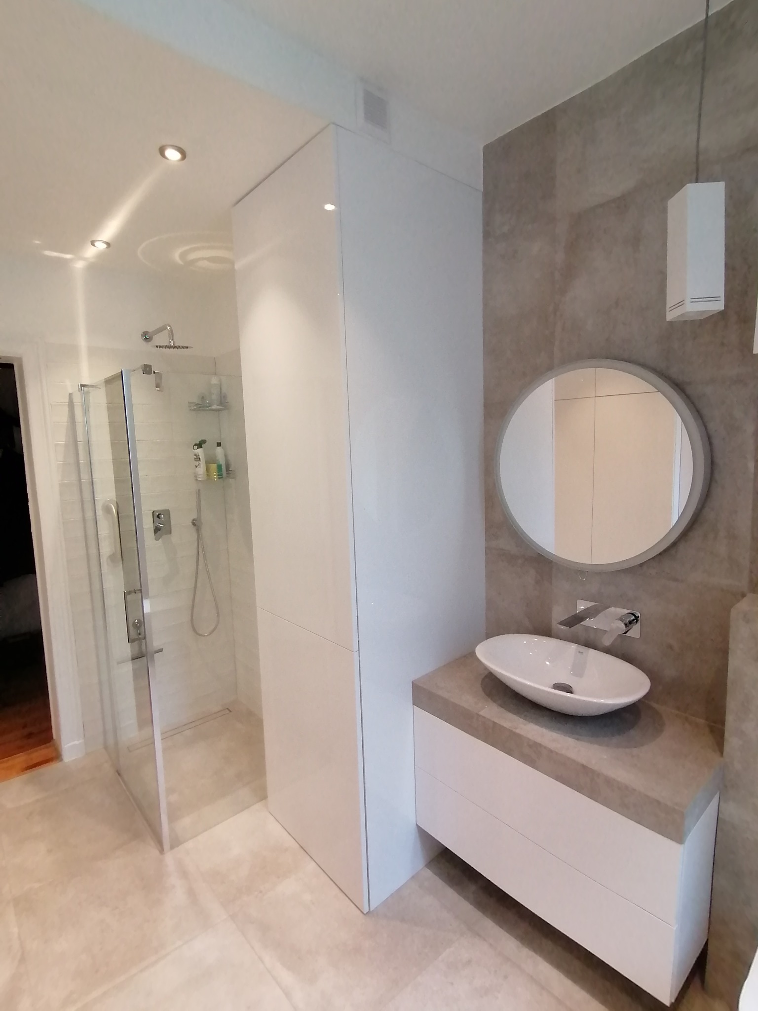 Nowoczesna łazienka z białą szafką pod umywalkę z szarym blatem, okrągłym lustrem i kabiną prysznicową typu walk-in z odpływem liniowym.