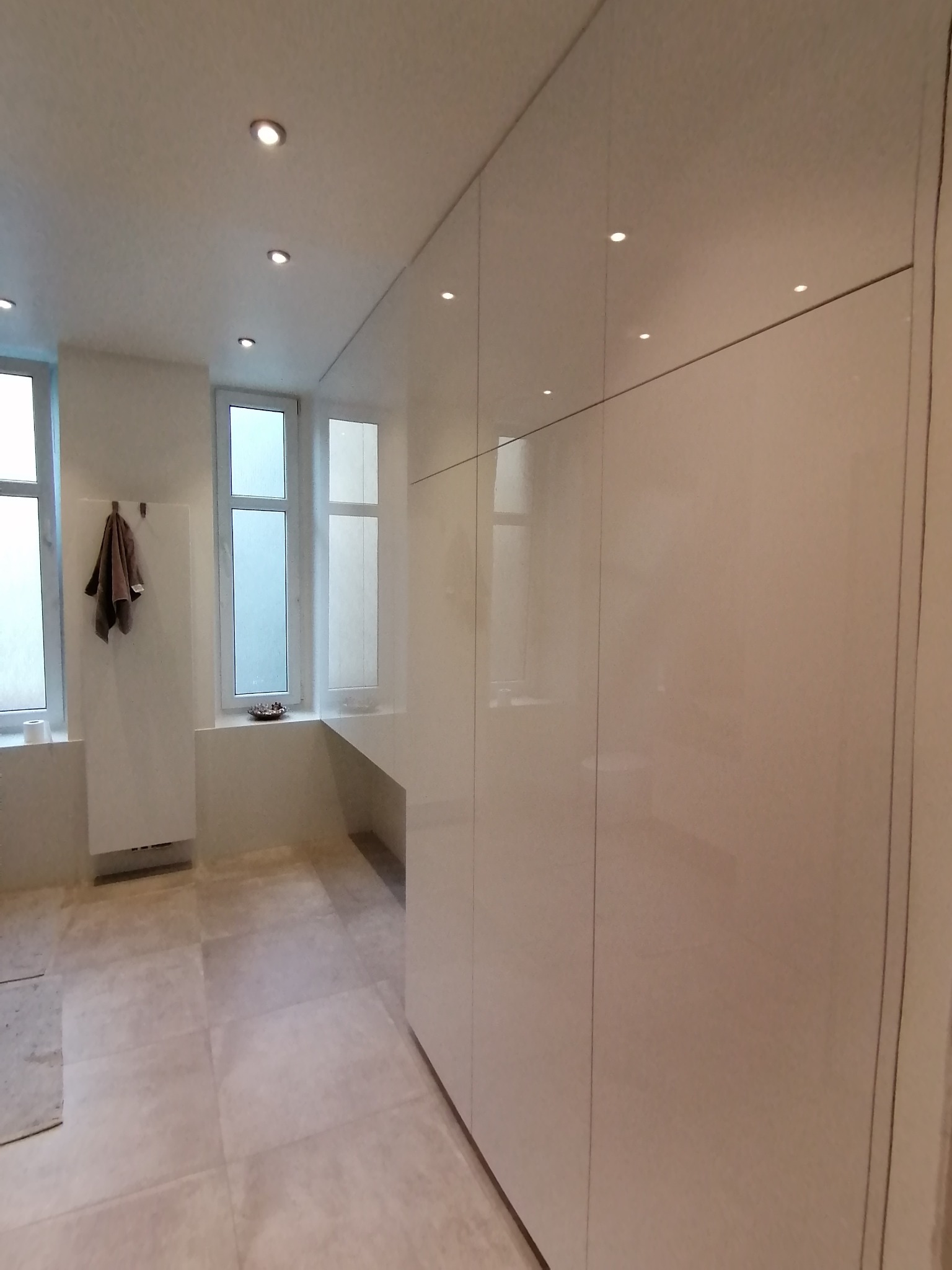 Biała, minimalistyczna zabudowa meblowa z połyskiem, zintegrowana ze ścianą, oświetlona punktowymi lampami sufitowymi, w tle okna z parapetem i grzejnik.