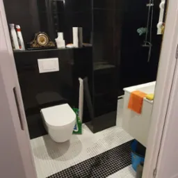 Nowoczesna łazienka z czarnymi płytkami na ścianach i białymi mozaikowymi płytkami na podłodze, widoczna toaleta, prysznic i akcesoria łazienkowe, żółta szmatka na podłodze przy progu.