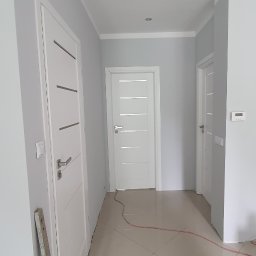 Coobmix - Korytarz z pomalowanymi na szaro ścianami, białe drzwi z poziomymi frezami, listwy przypodłogowe, płytki podłogowe w odcieniach beżu, widoczny kabel i wiadro z resztkami farby.