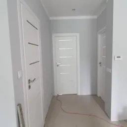 Korytarz z pomalowanymi na szaro ścianami, białe drzwi z poziomymi frezami, listwy przypodłogowe, płytki podłogowe w odcieniach beżu, widoczny kabel i wiadro z resztkami farby.