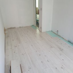 Coobmix - Układanie jasnej podłogi laminowanej w pomieszczeniu z białymi ścianami, widoczna warstwa izolacyjna i przygotowane panele.