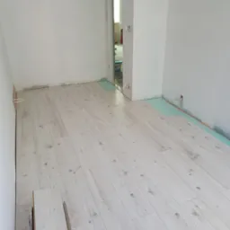 Układanie jasnej podłogi laminowanej w pomieszczeniu z białymi ścianami, widoczna warstwa izolacyjna i przygotowane panele.