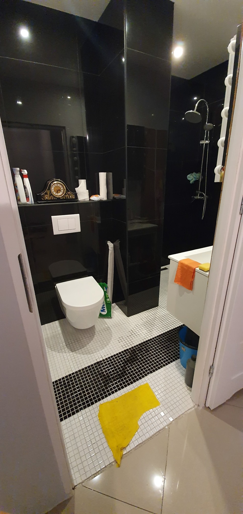 Nowoczesna łazienka z czarnymi płytkami na ścianach i białymi mozaikowymi płytkami na podłodze, widoczna toaleta, prysznic i akcesoria łazienkowe, żółta szmatka na podłodze przy progu.