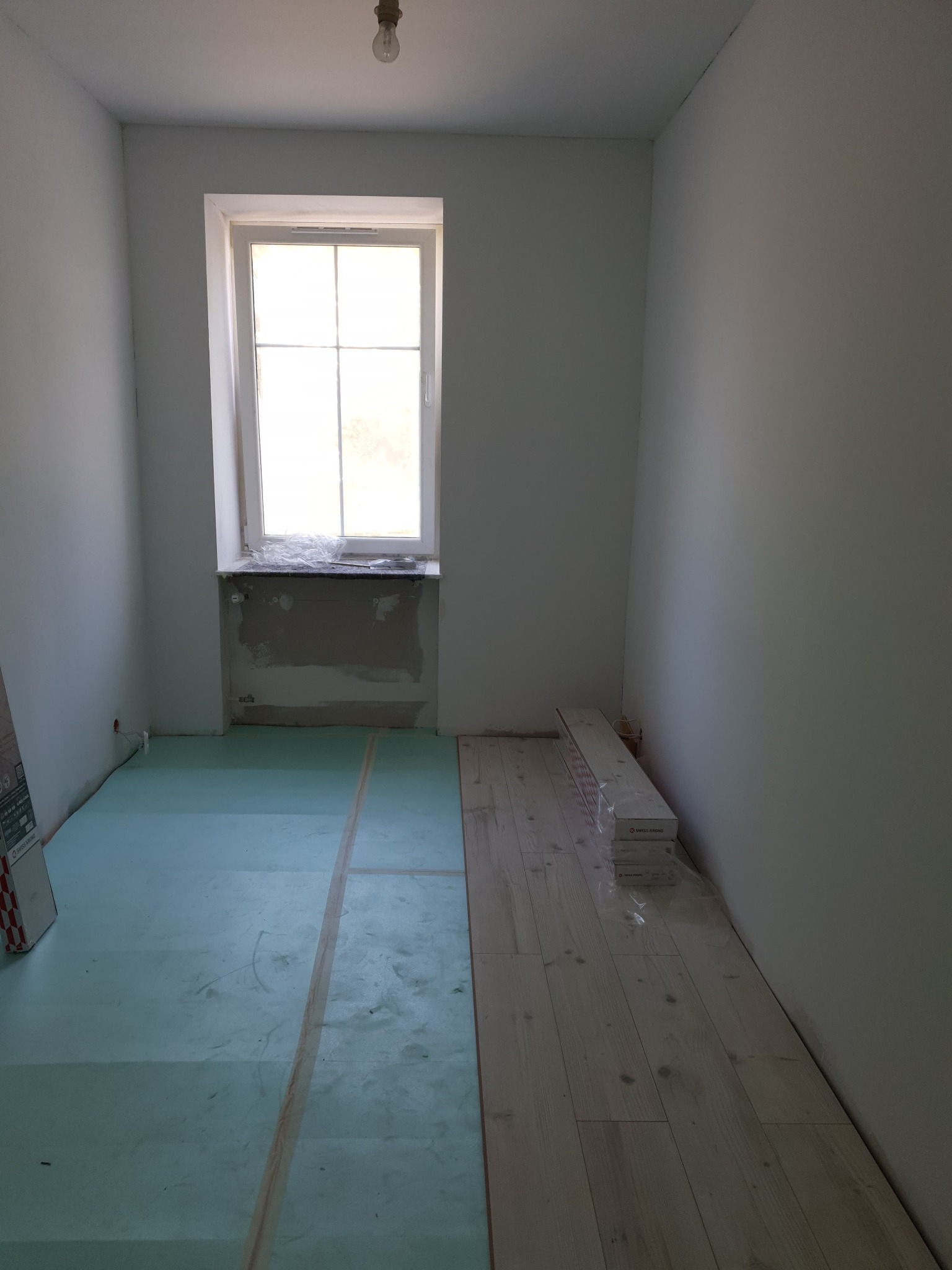 Pokój w trakcie układania podłogi: część z podkładem izolacyjnym, część z ułożonymi jasnymi panelami, paczki paneli przy ścianie, przygotowane podłoże pod oknem.