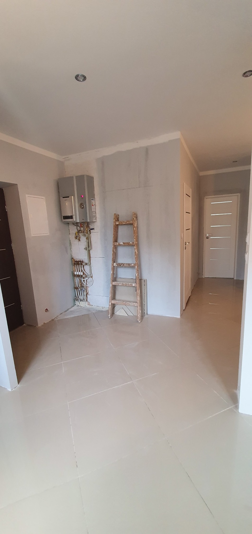Wnętrze w trakcie prac wykończeniowych: położone płytki podłogowe, drabina oparta o ścianę z instalacją grzewczą, widoczny bojler i przygotowane ściany pod malowanie.