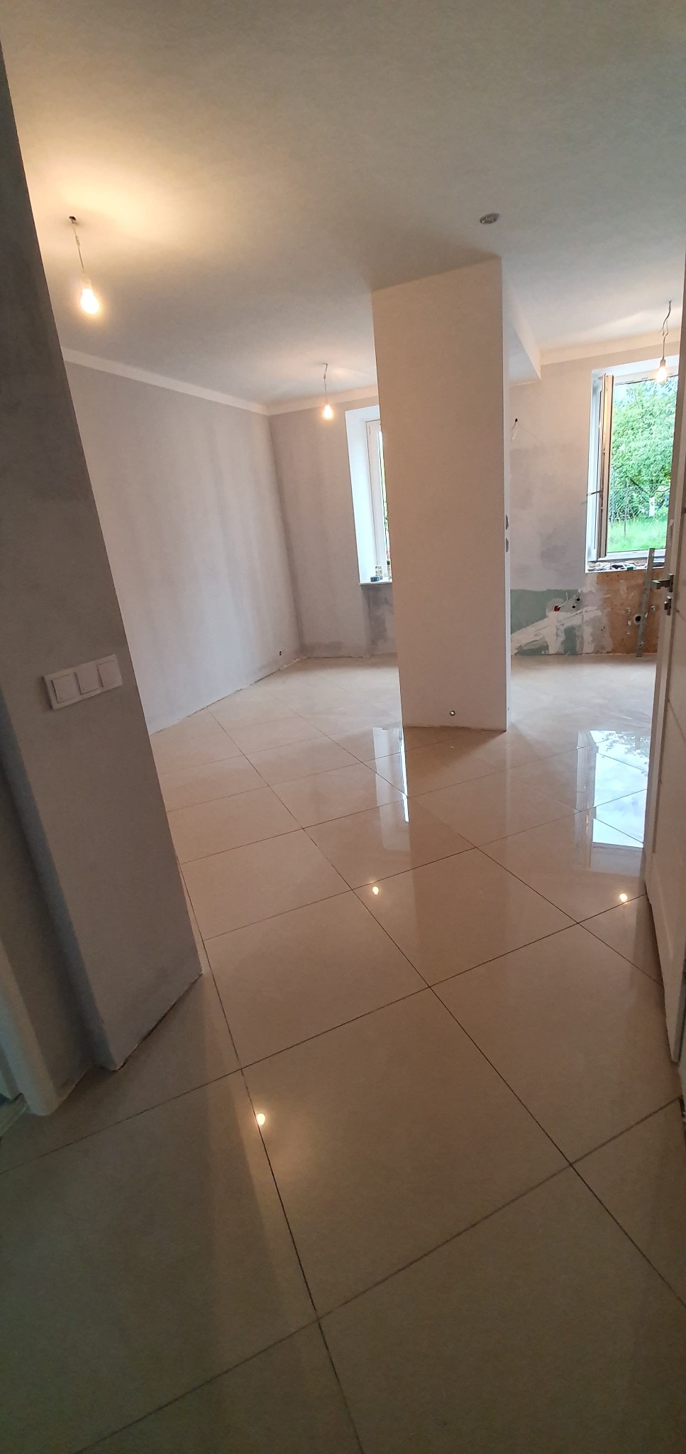 Wnętrze w trakcie remontu: beżowe płytki podłogowe z połyskiem, surowe ściany z widocznymi śladami prac wykończeniowych, okna z widokiem na zieleń, wiszące żarówki bez kloszy.