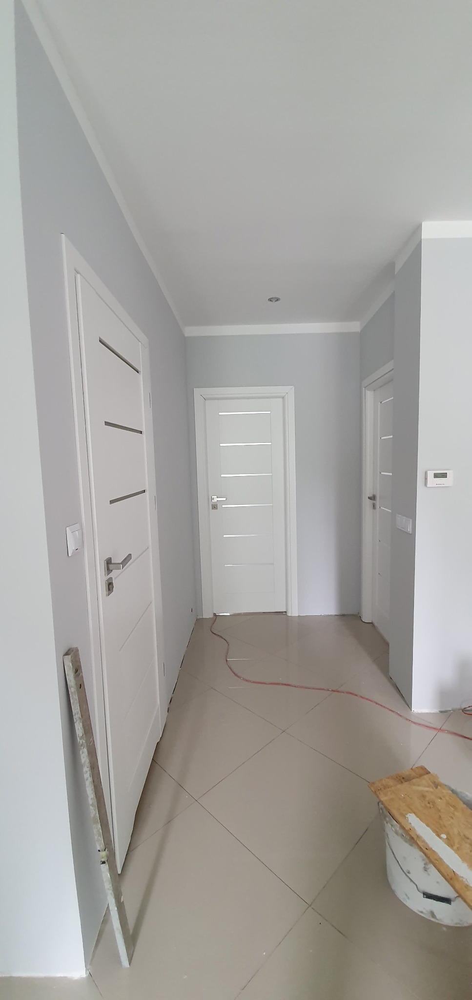 Korytarz z pomalowanymi na szaro ścianami, białe drzwi z poziomymi frezami, listwy przypodłogowe, płytki podłogowe w odcieniach beżu, widoczny kabel i wiadro z resztkami farby.