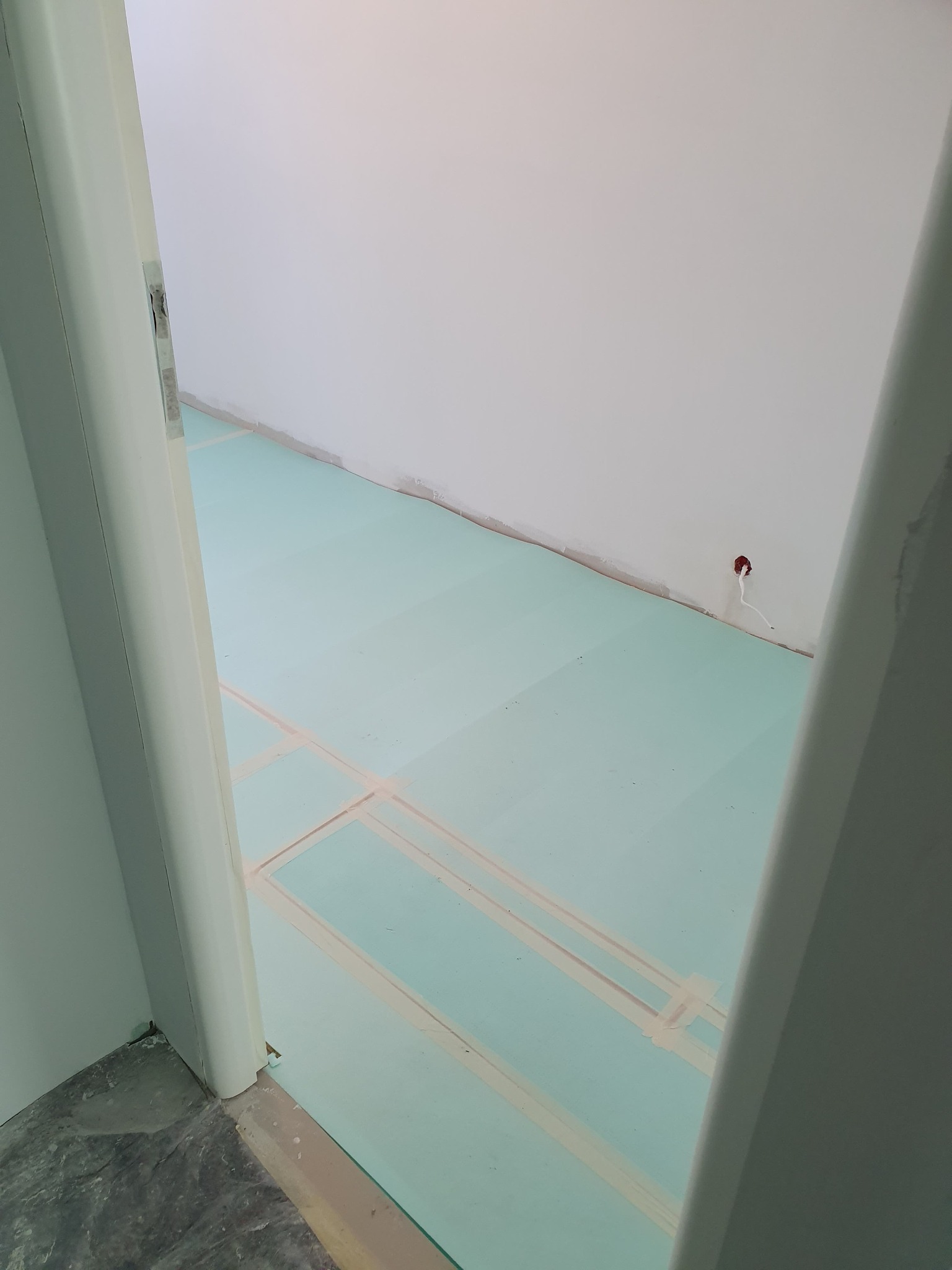 Przygotowanie podłoża pod podłogę: płyty izolacyjne z taśmą na wylewce, widok zza futryny drzwiowej.