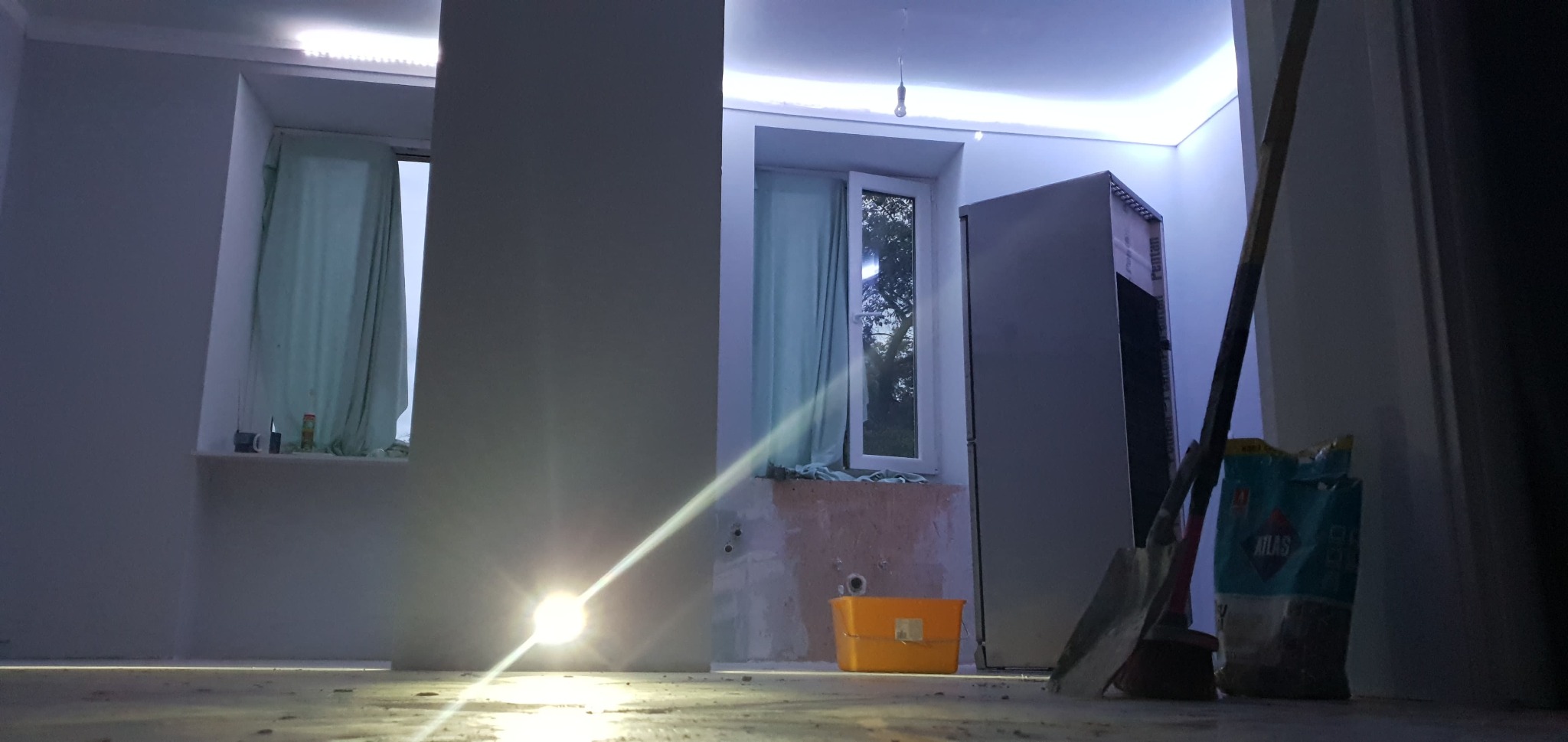 Wnętrze pokoju podczas prac remontowych: widoczne dwa okna z zasłonami, fragment ściany z instalacją, wiadro, szpachla i worek zaprawy.