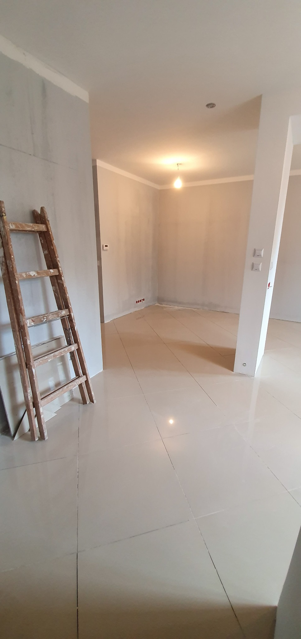 Wnętrze w trakcie prac wykończeniowych: jasne płytki podłogowe, surowe ściany z listwami, drewniana drabina oparta o ścianę, widoczne gniazdka elektryczne.