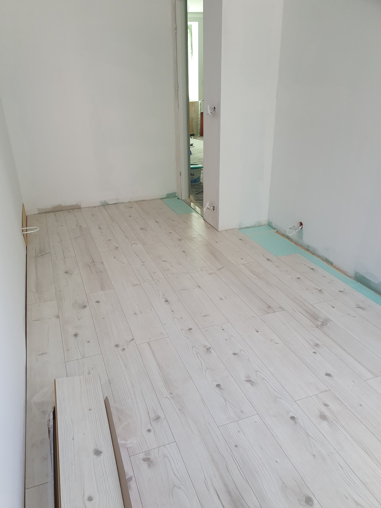 Układanie jasnej podłogi laminowanej w pomieszczeniu z białymi ścianami, widoczna warstwa izolacyjna i przygotowane panele.