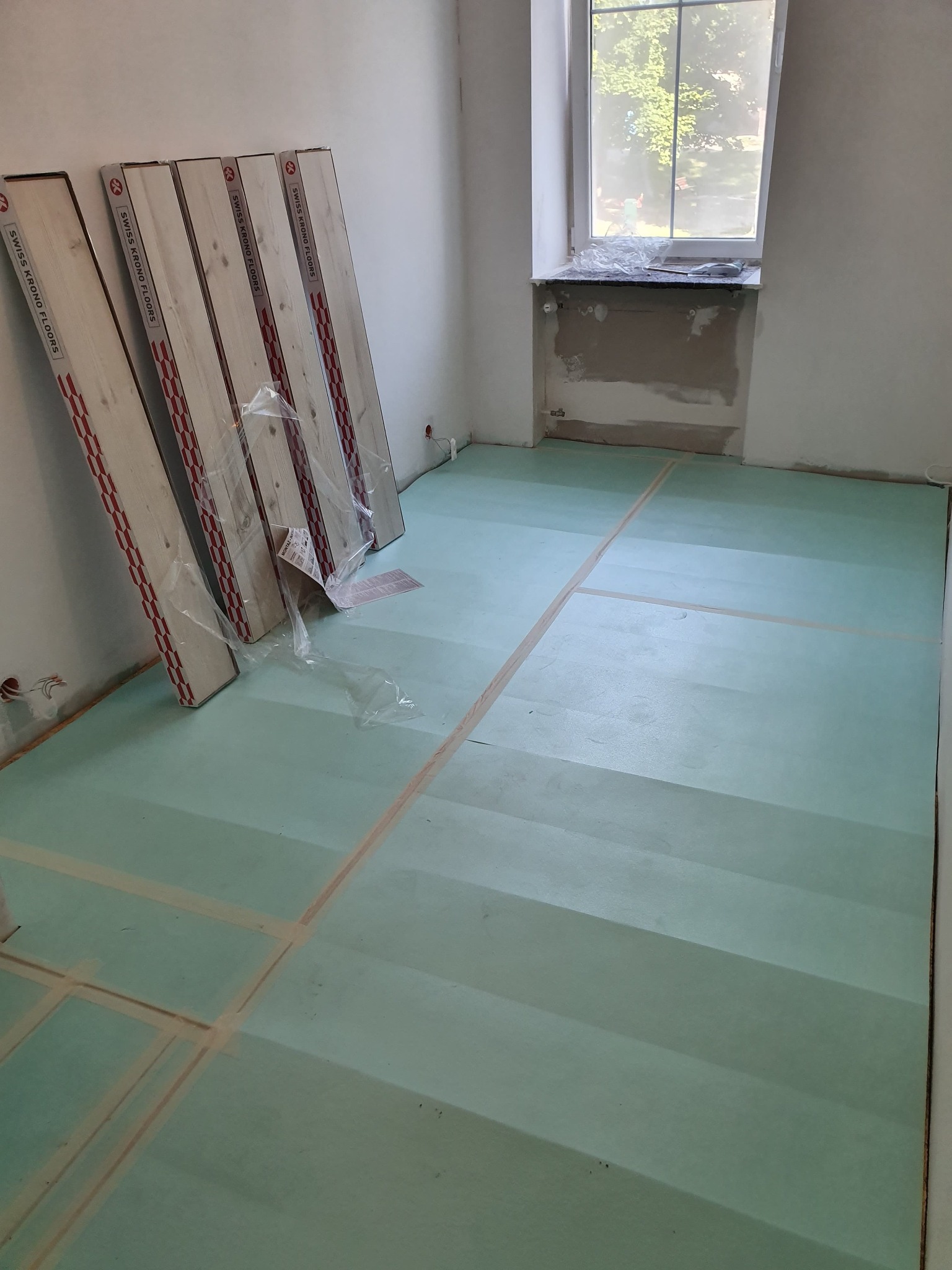 Przygotowanie do montażu podłogi: rozłożona zielona pianka izolacyjna z taśmą wyznaczającą obszary, oraz paczki paneli Swiss Krono Floors o jasnym, drewnopodobnym wzorze, opartych o ścianę w pustym...