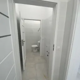 Nowoczesna, minimalistyczna łazienka z białymi płytkami, widoczna przez otwarte drzwi, z toaletą w tle i szarymi płytkami podłogowymi.