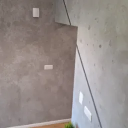 Szara ściana z efektem betonu architektonicznego, oddzielona czarną listwą od drugiej ściany, drewniana podłoga i szafka nocna z roślinką doniczkową i zegarkiem.