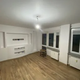 Pusty pokój po remoncie z drewnianą podłogą, białymi ścianami z wnękami i grzejnikami pod oknami. Widoczny stolik i wisząca żarówka.