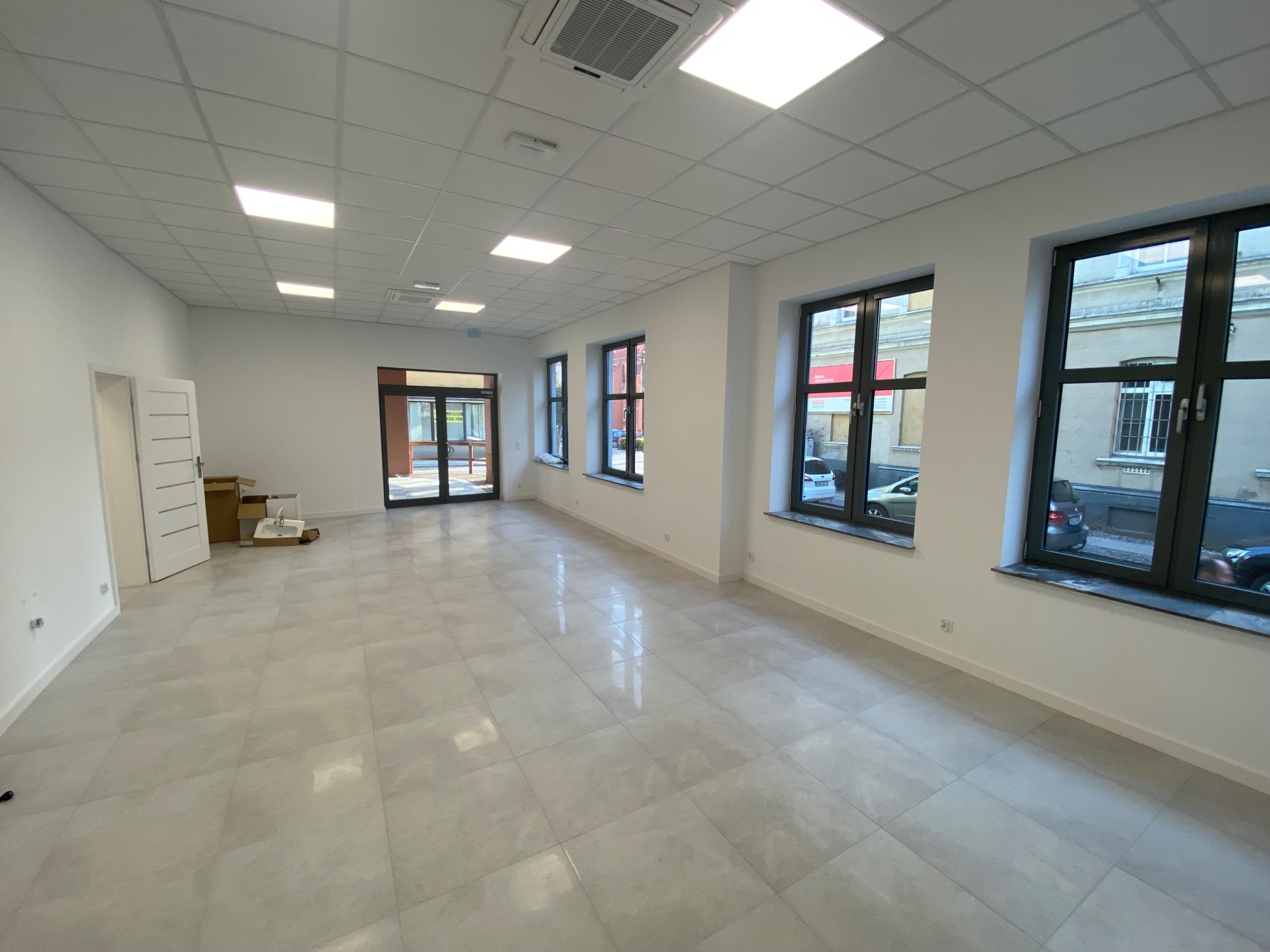 Jasne, przestronne wnętrze biurowe z nową, błyszczącą podłogą z płytek, białymi ścianami, dużymi oknami w ciemnych ramach i podwieszanym sufitem z oświetleniem LED.
