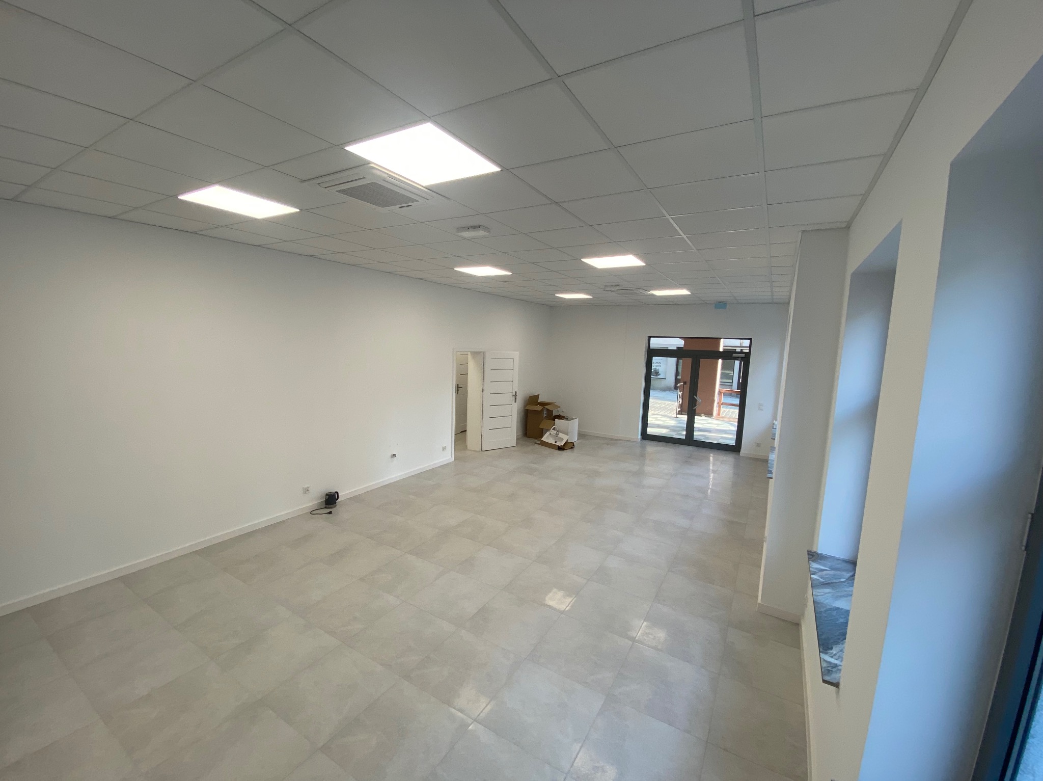 Wnętrze nowo wykończonego, przestronnego pomieszczenia z jasnymi płytkami podłogowymi, białymi ścianami, podwieszanym sufitem z panelami LED i wnęką okienną wykończoną marmurem.