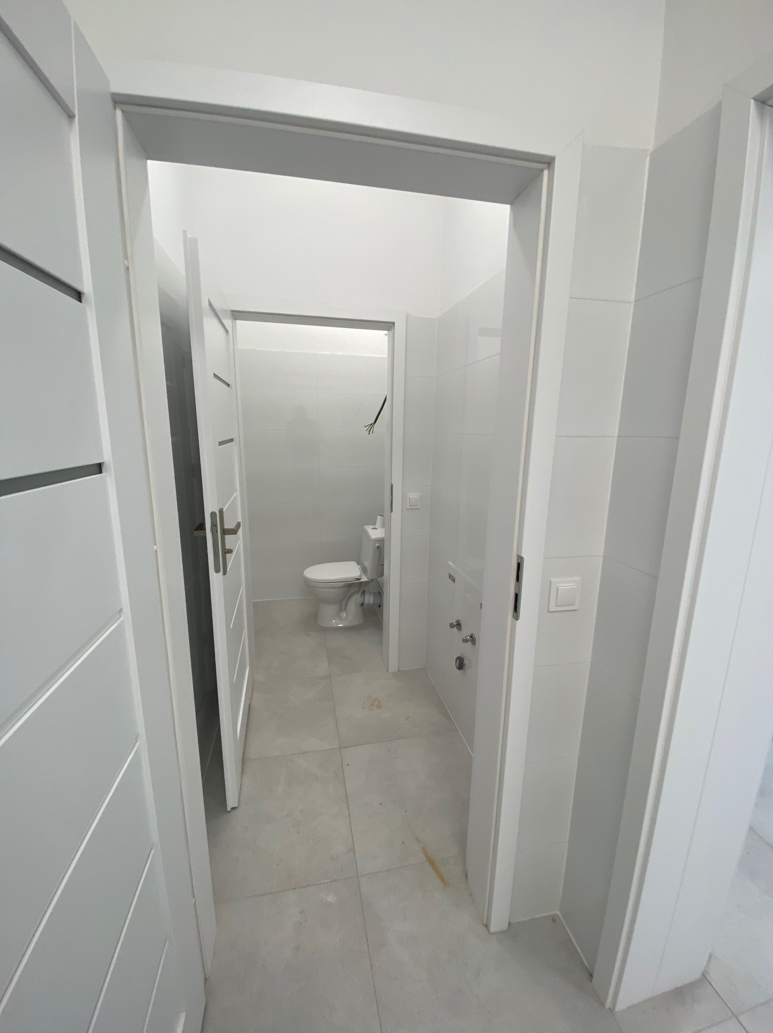 Nowoczesna, minimalistyczna łazienka z białymi płytkami, widoczna przez otwarte drzwi, z toaletą w tle i szarymi płytkami podłogowymi.