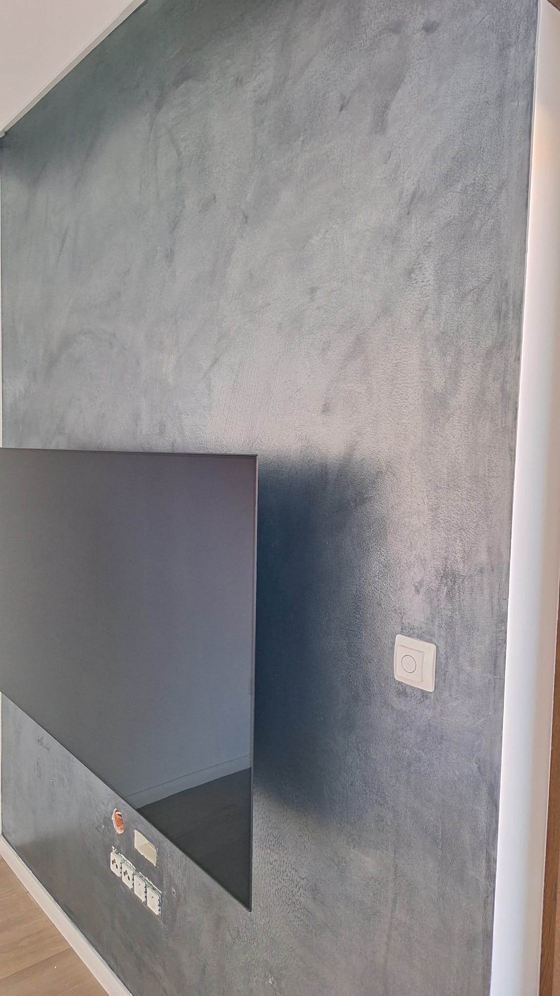 Ściana pokryta dekoracyjną, szarą farbą z efektem metalicznego połysku, widoczne gniazdka elektryczne i włącznik światła.