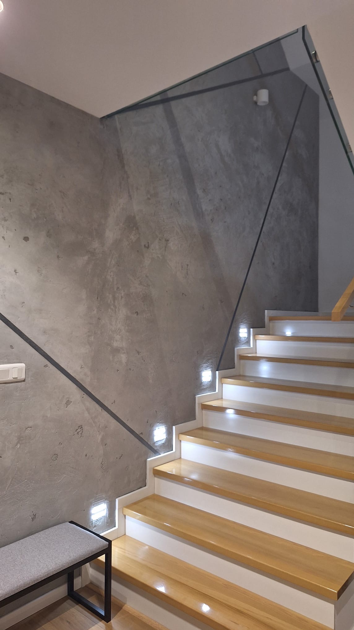 Schody drewniane z białymi podstopnicami, oświetleniem LED w ścianie pokrytej szarą gładzią dekoracyjną i szklaną balustradą.