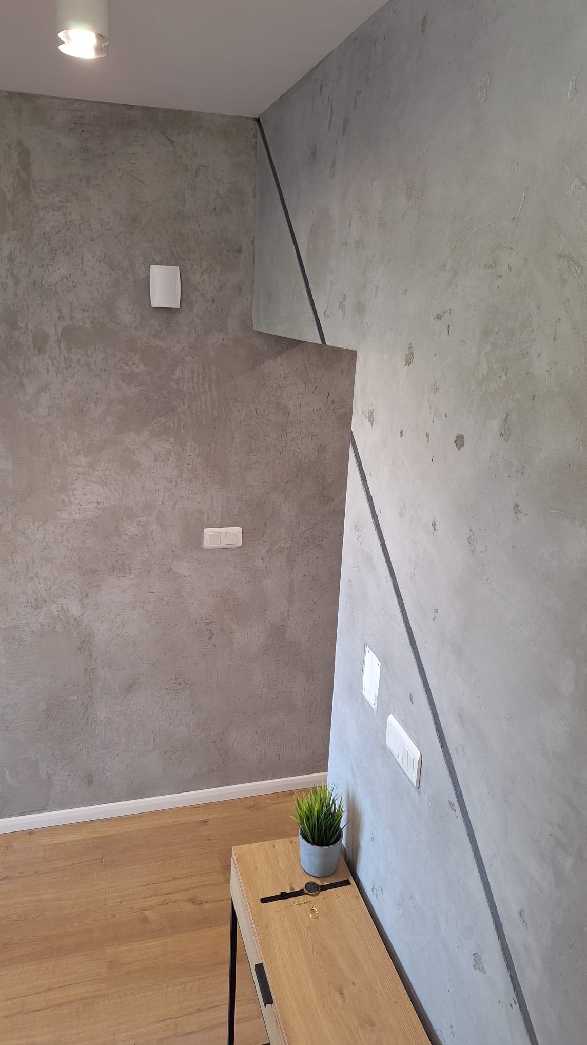 Szara ściana z efektem betonu architektonicznego, oddzielona czarną listwą od drugiej ściany, drewniana podłoga i szafka nocna z roślinką doniczkową i zegarkiem.