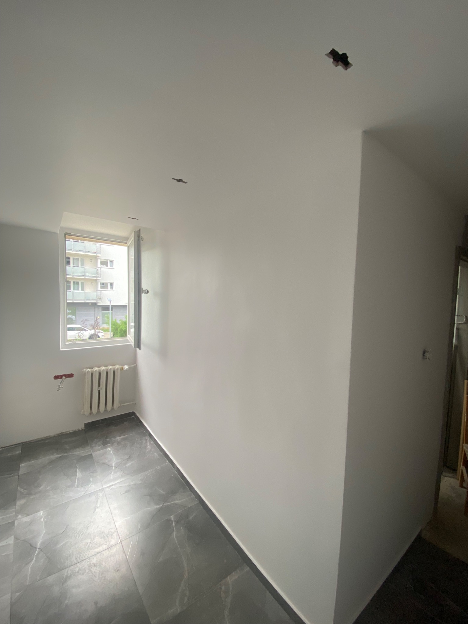 Pokój po świeżym malowaniu ścian na biało, z nową podłogą z szarych płytek imitujących marmur, widoczne przygotowania do montażu oświetlenia sufitowego.