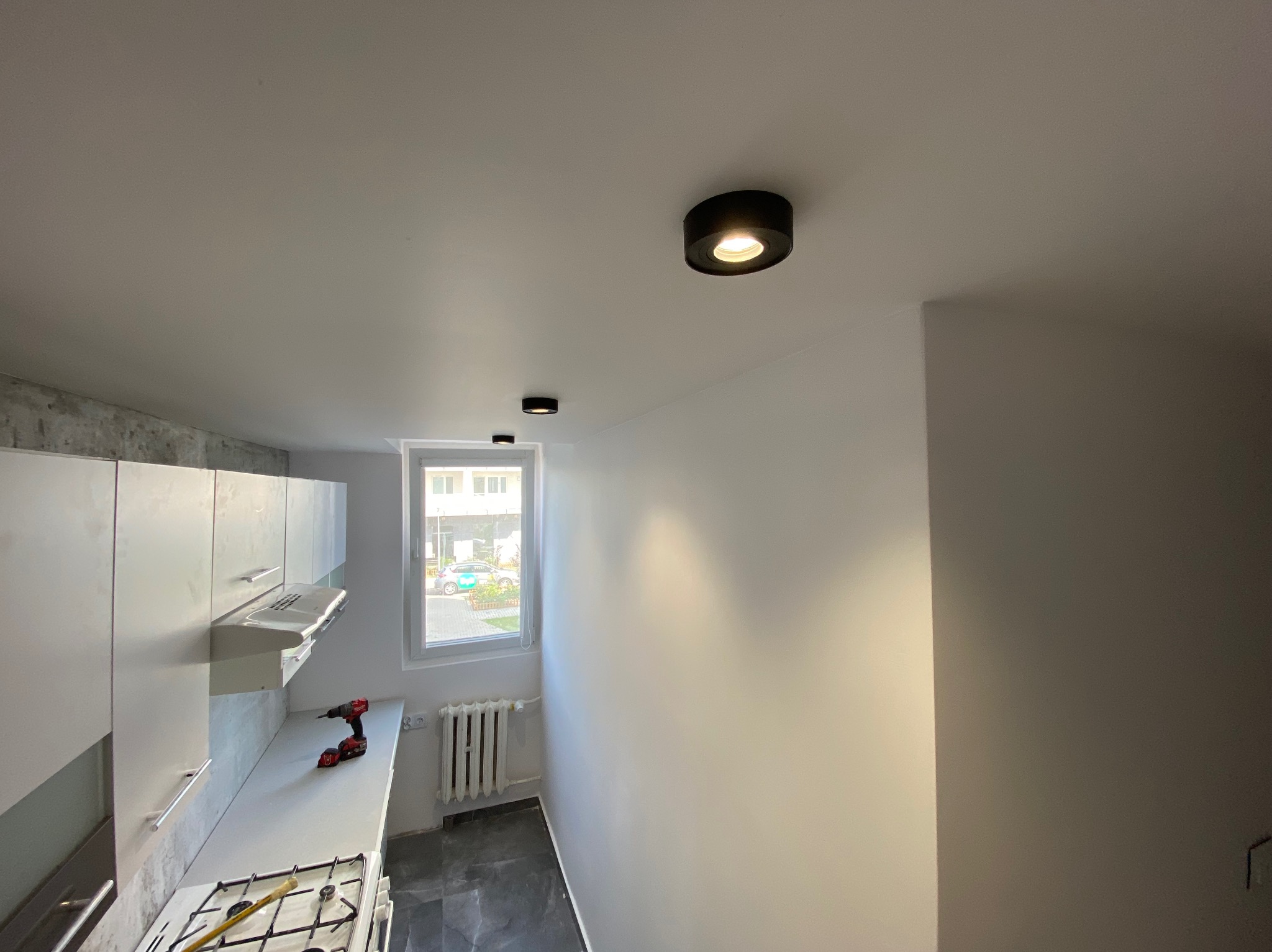 Kuchnia po remoncie z białymi szafkami, marmurowym blatem, czarnymi, okrągłymi lampami sufitowymi i narzędziami leżącymi na blacie.