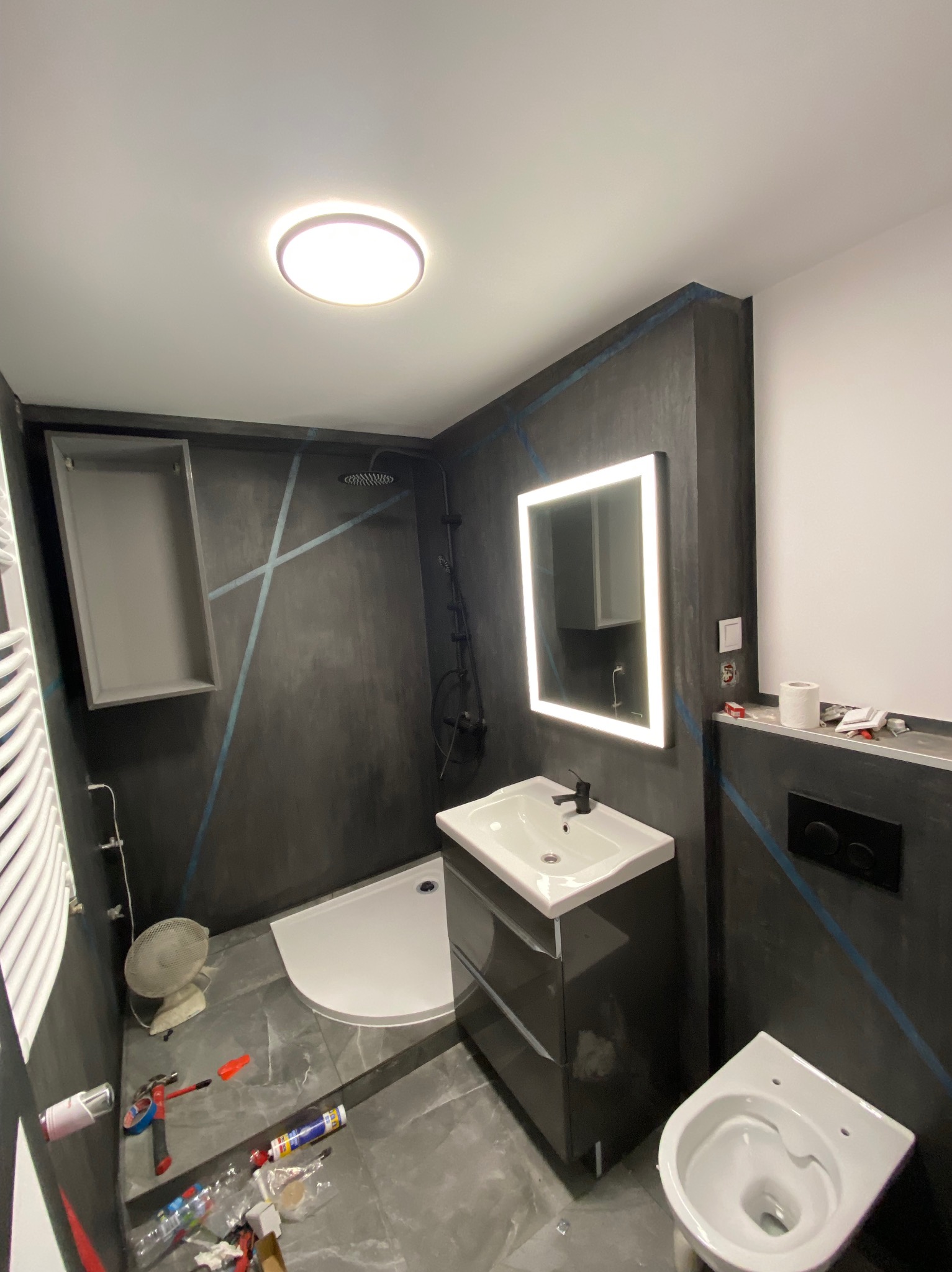Ciemna łazienka w trakcie prac wykończeniowych, widoczna kabina prysznicowa, umywalka z szafką, toaleta oraz narzędzia i materiały budowlane na podłodze.