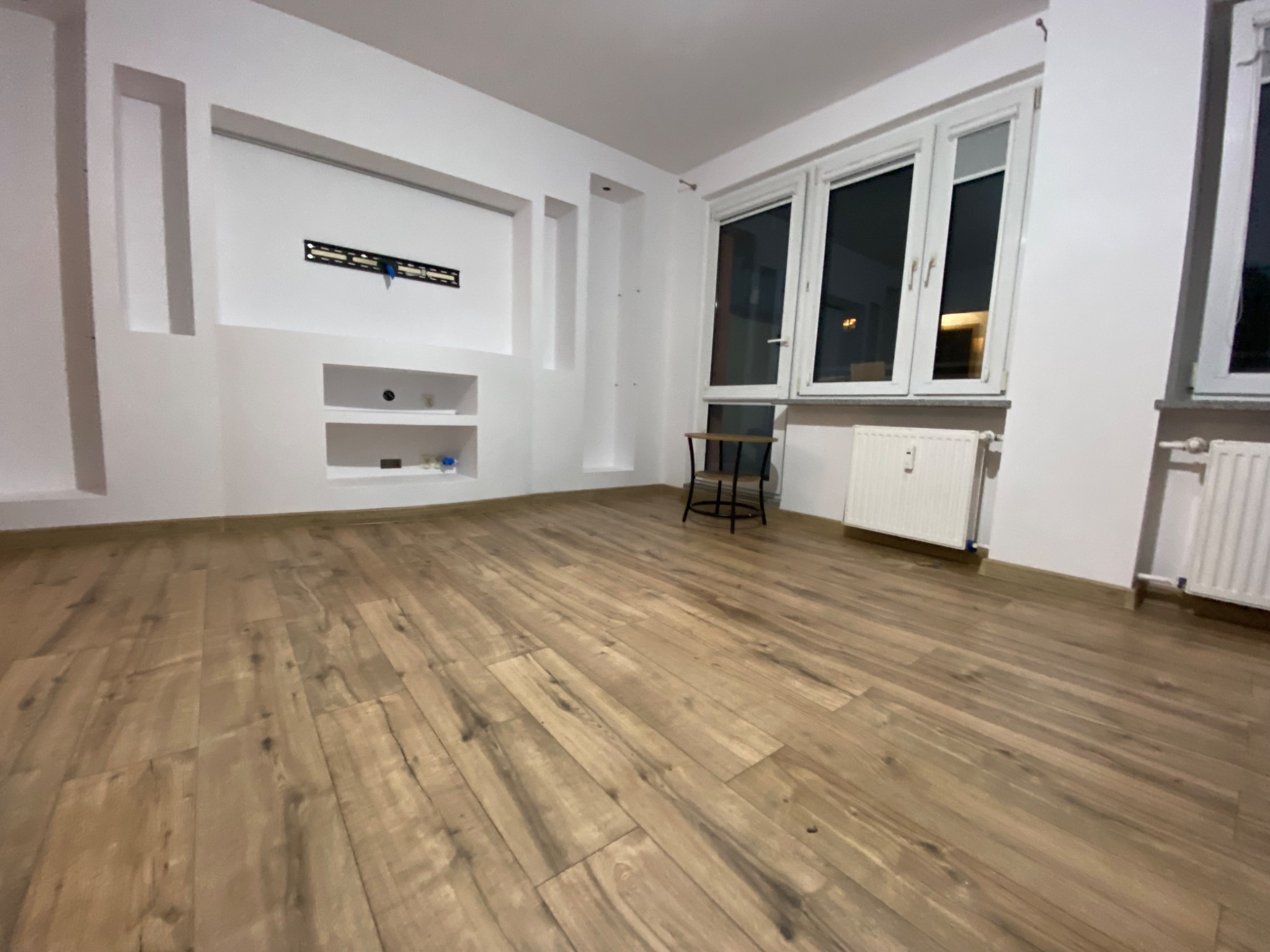 Pusty pokój z drewnianą podłogą, białe ściany z wnękami, zamontowany uchwyt na telewizor, okna z widokiem na nocne miasto, grzejniki pod oknami, mały stolik.