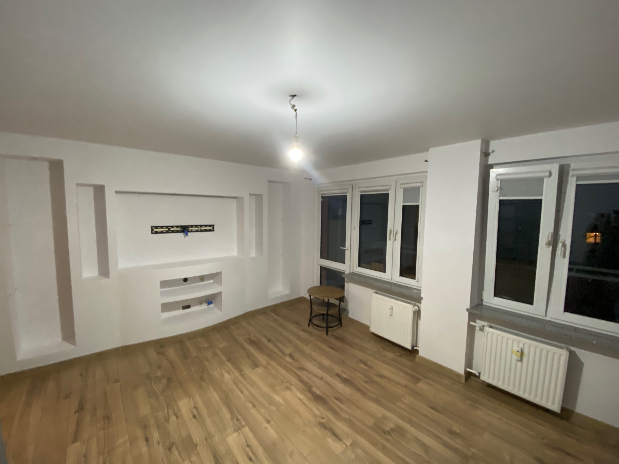 Pusty pokój po remoncie z drewnianą podłogą, białymi ścianami z wnękami i grzejnikami pod oknami. Widoczny stolik i wisząca żarówka.