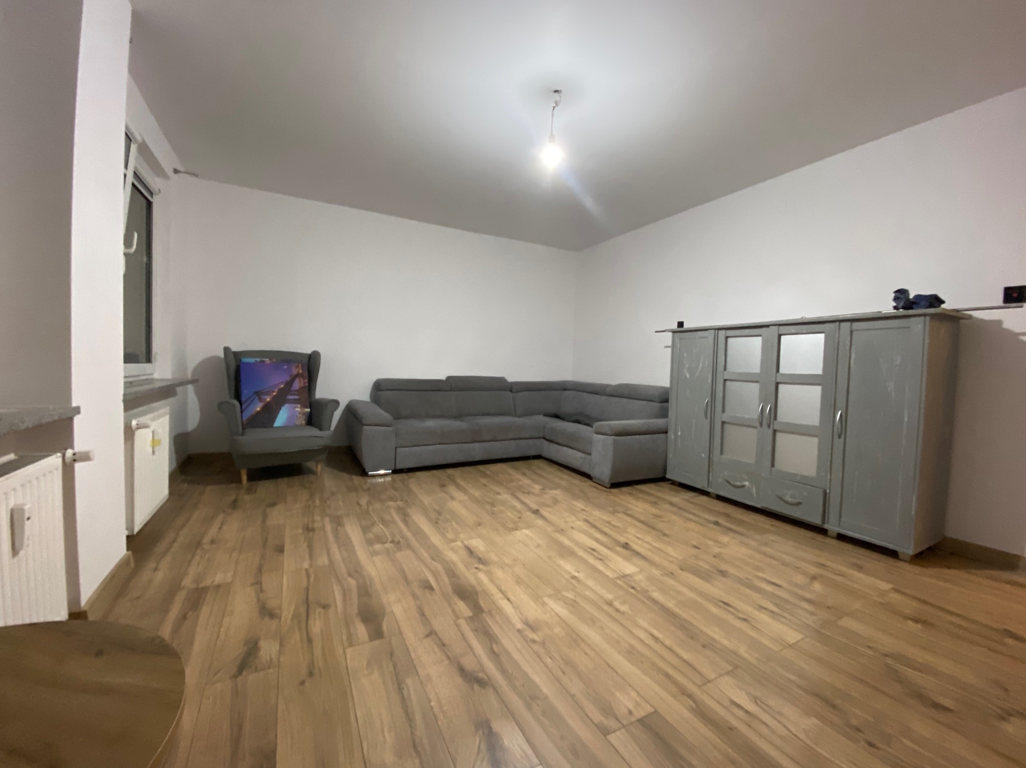 Jasne wnętrze pokoju z drewnianą podłogą, szarą narożną sofą, fotelem z obrazem na oparciu i szarą komodą z przeszkleniami.