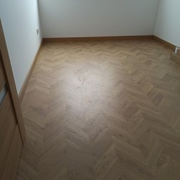 PAW-ZONE Prace Wykonczniowe - Ukończona podłoga z paneli ułożonych we wzór jodełki w jasnym pokoju z białymi ścianami i oknem zabezpieczonym folią malarską.