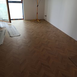 PAW-ZONE Prace Wykonczniowe - Ułożona, wzorzysta podłoga drewniana w jodełkę, z widocznymi kartonami po panelach i lampą budowlaną w pustym pomieszczeniu z oknem.