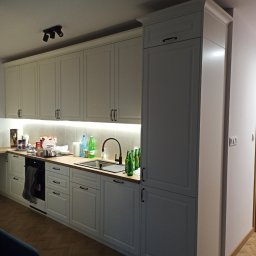 PAW-ZONE Prace Wykonczniowe - Biała zabudowa kuchenna z oświetleniem podszafkowym, czarną armaturą i panelami podłogowymi ułożonymi w jodełkę; na blacie widoczne butelki i akcesoria kuchenne.