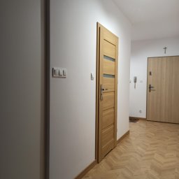 PAW-ZONE Prace Wykonczniowe - Jasny korytarz po remoncie z parkietem w jodełkę, dwiema parami drzwi z przeszkleniami i gładkimi, białymi ścianami.