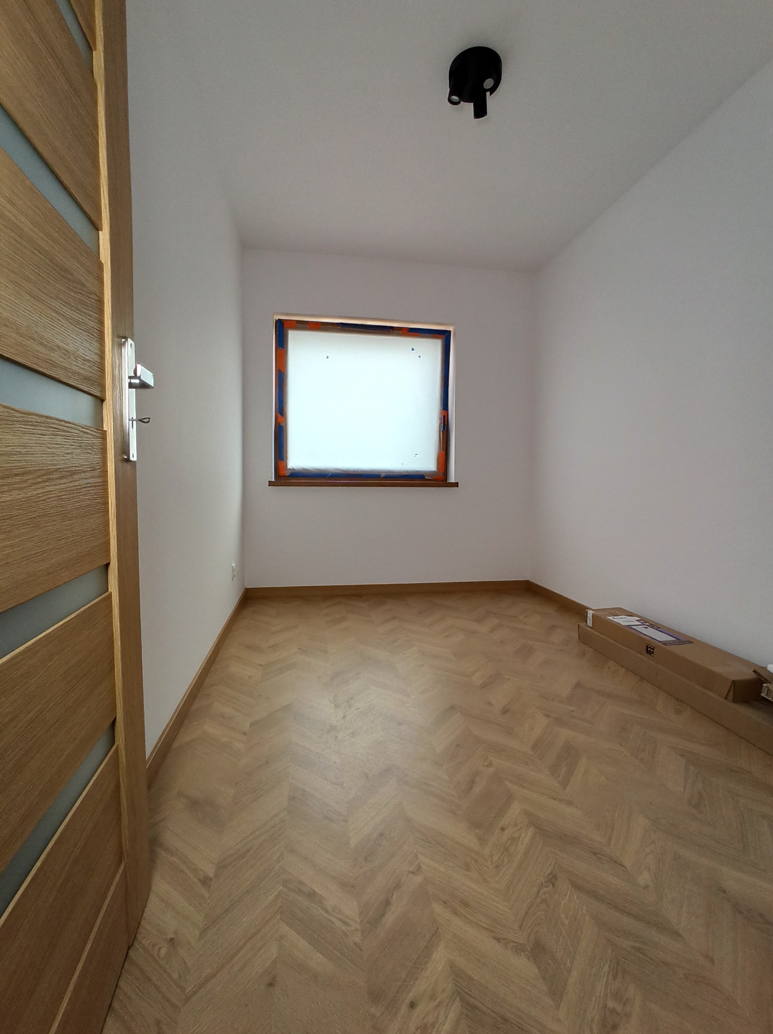 Pusty pokój po remoncie z parkietem ułożonym w jodełkę, białe ściany, okno zabezpieczone folią malarską i drewniane drzwi z przeszkleniami.