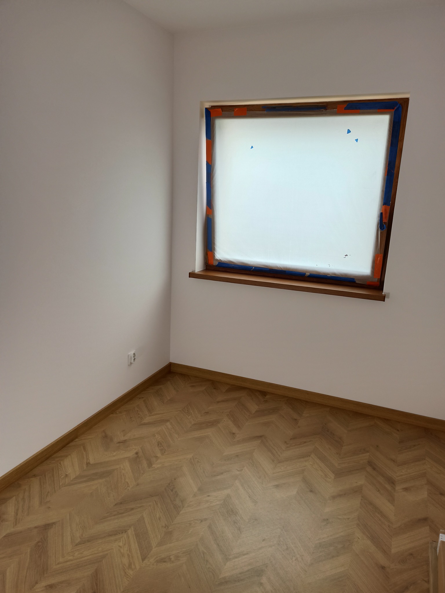 Pokój przed malowaniem: zabezpieczone okno folią i taśmą, świeżo położona drewniana podłoga w jodełkę, gładkie białe ściany.