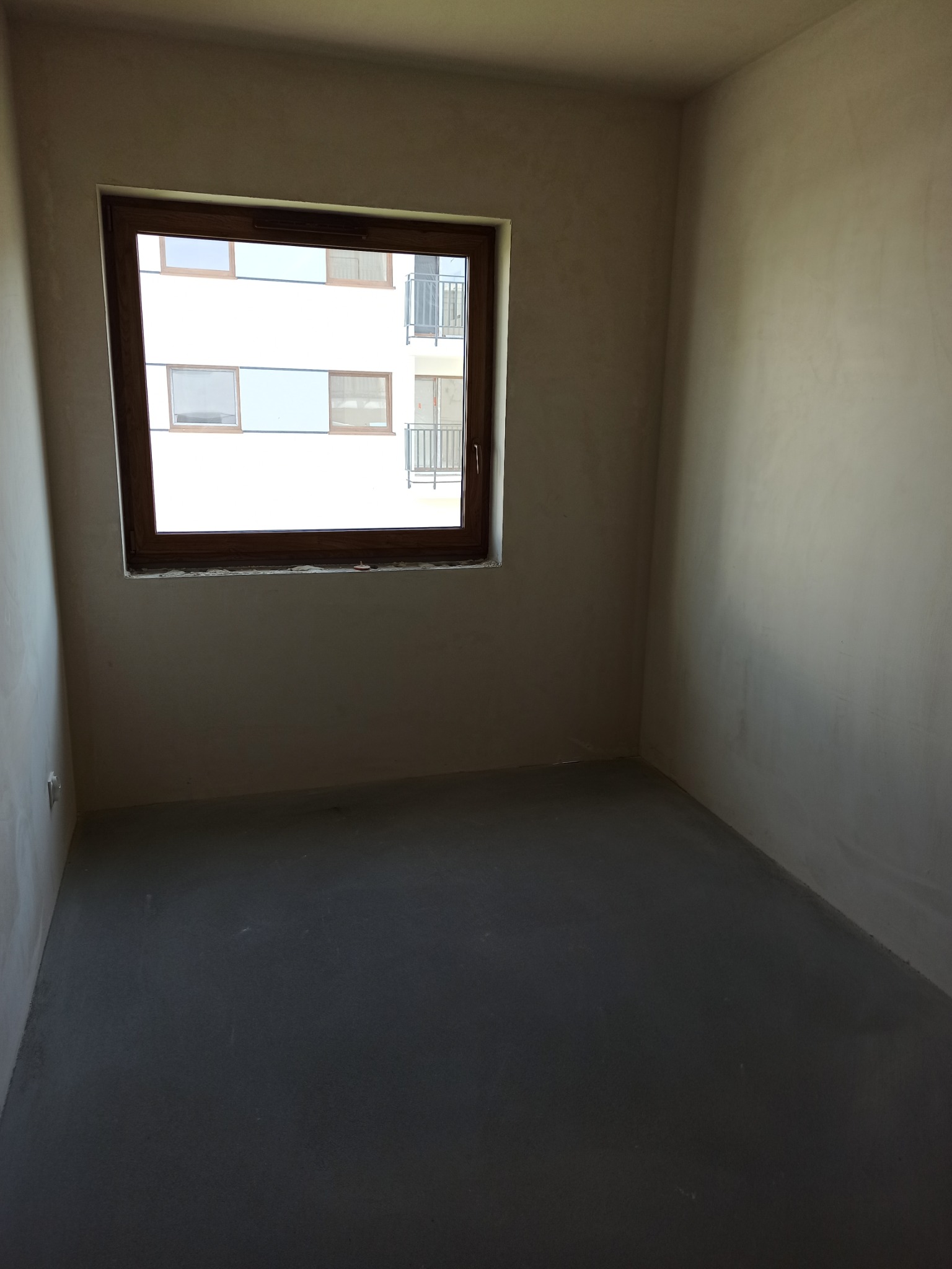 Surowe wnętrze pokoju z oknem, ściany pokryte świeżą warstwą szpachli, podłoga wylewka samopoziomująca, widok z okna na biały budynek z balkonami.