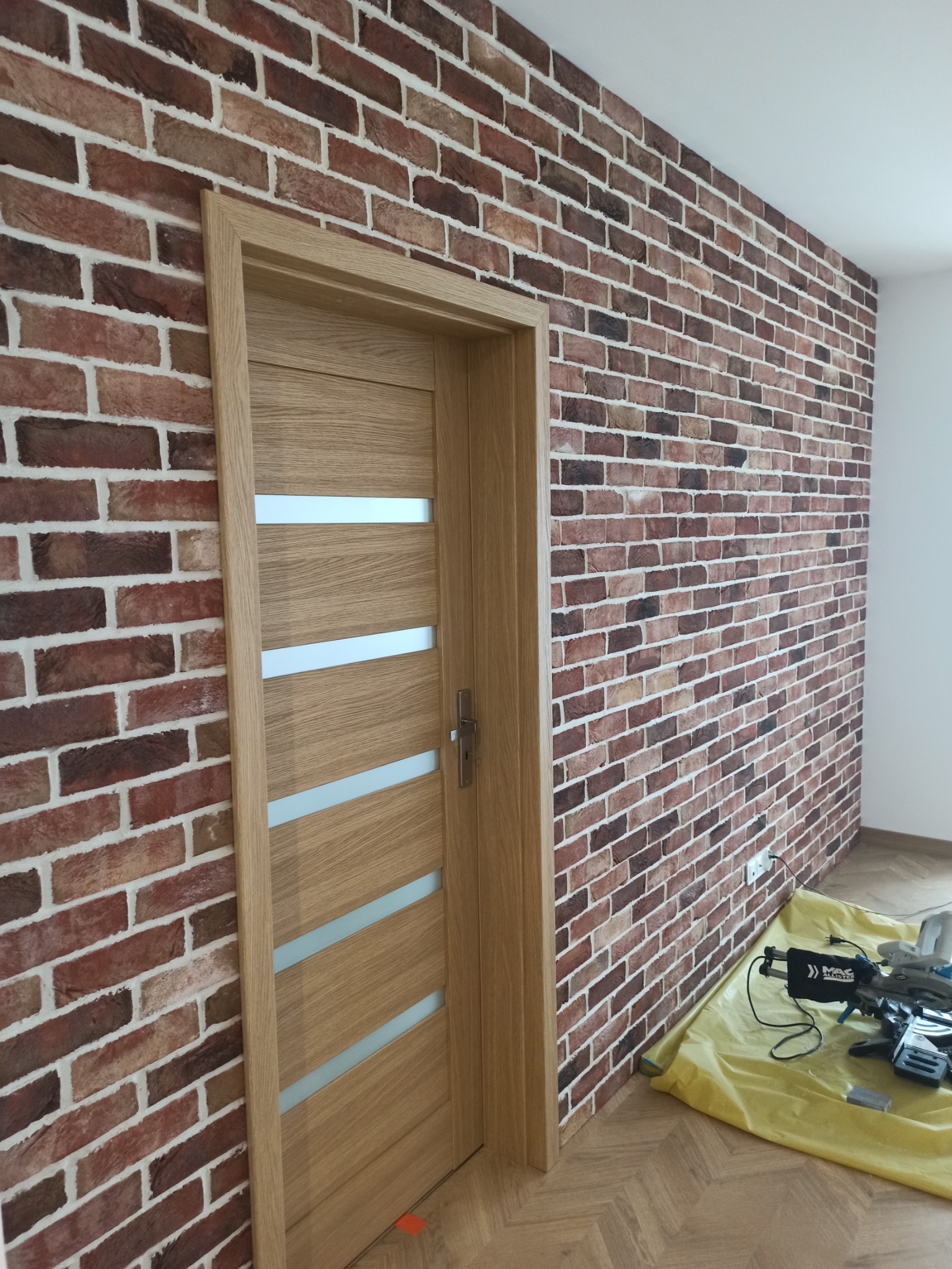 Dębowa ościeżnica z zamontowanymi drzwiami z przeszkleniami na tle ściany z cegły klinkierowej, widoczne narzędzia na podłodze zabezpieczonej folią.