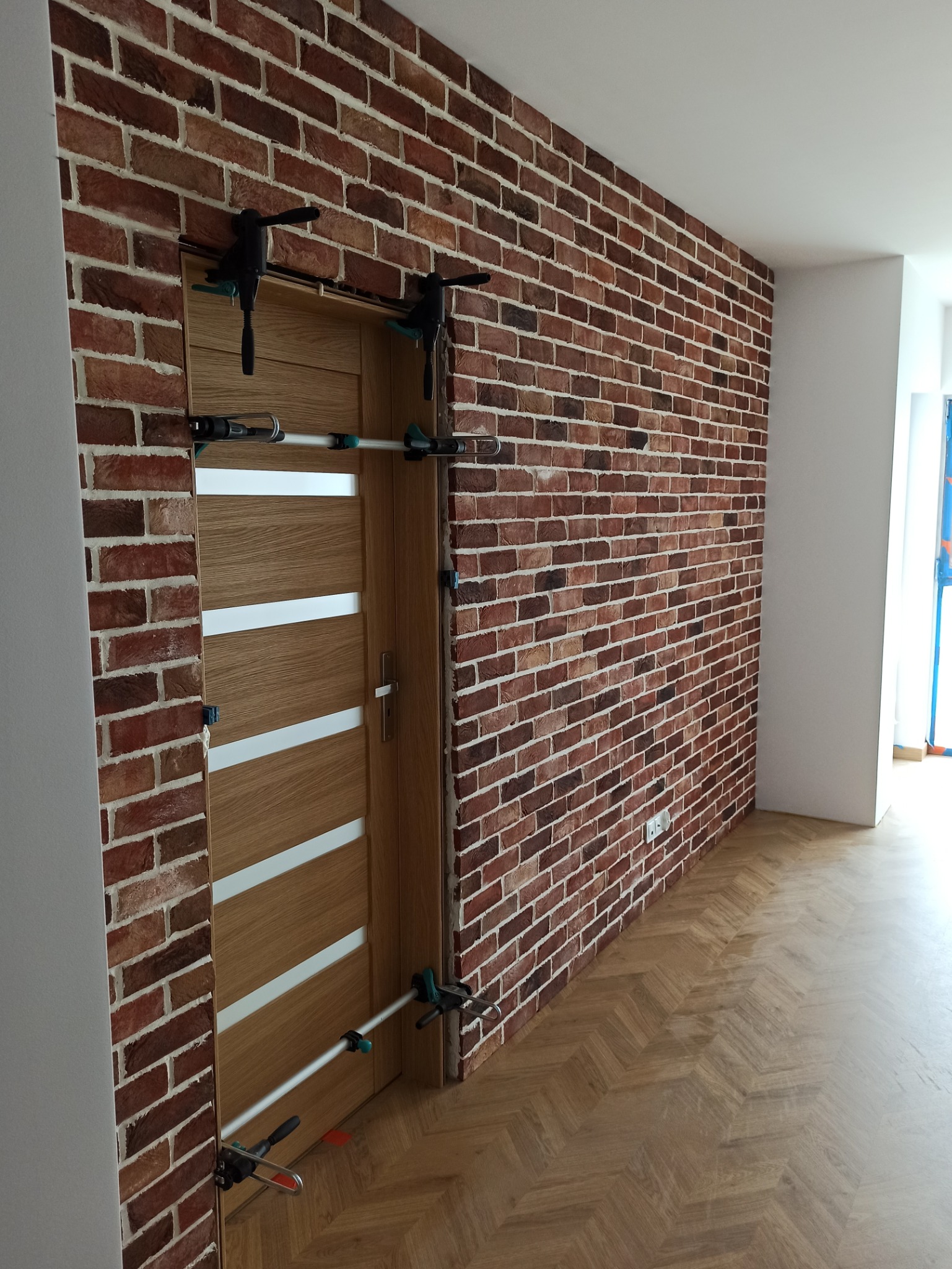 Montaż drzwi z drewnianą okleiną i białymi paskami, przytrzymywanych przez ściski stolarskie na tle ściany wykończonej cegłą klinkierową i parkietu w jodełkę.