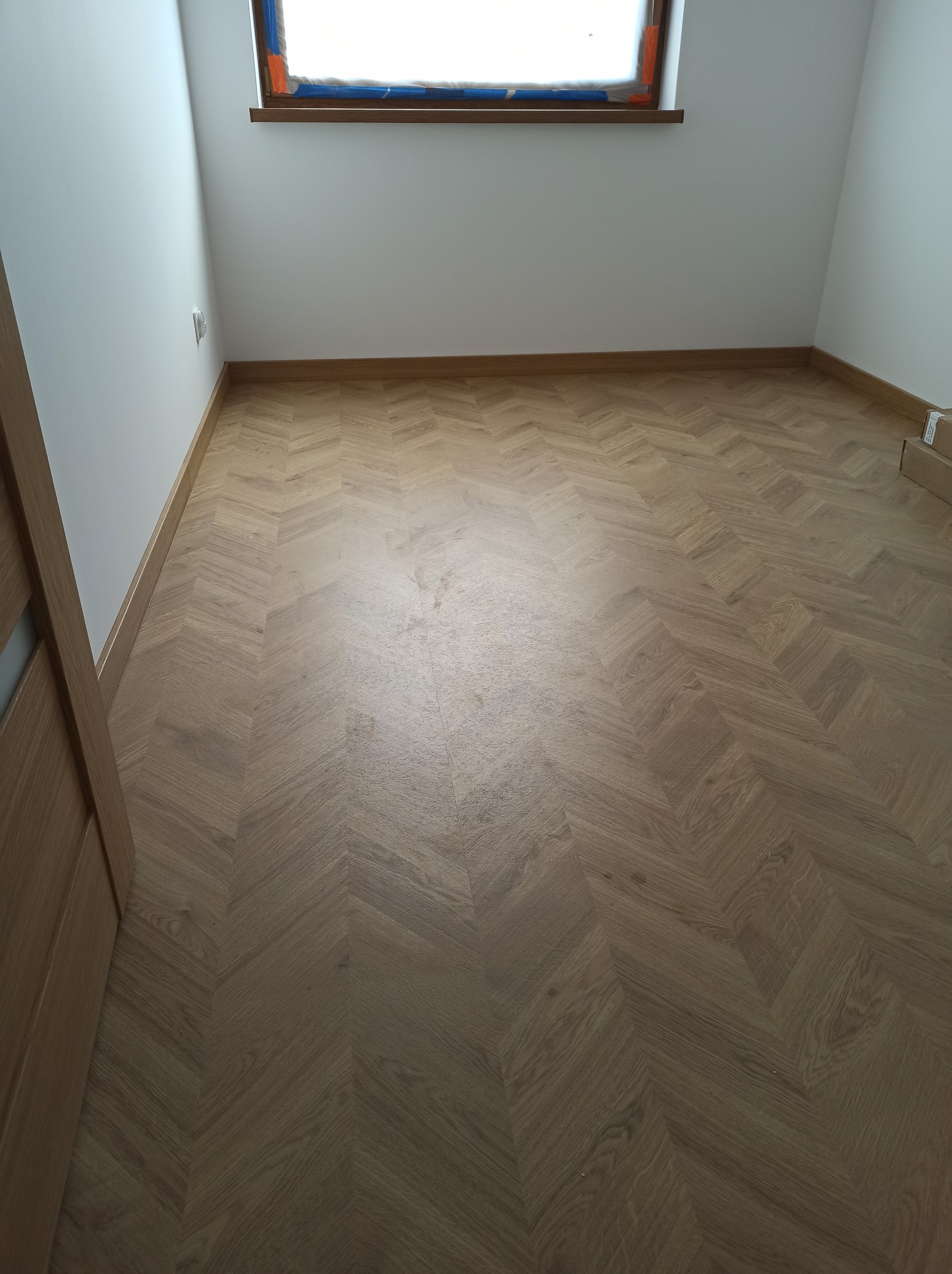Ukończona podłoga z paneli ułożonych we wzór jodełki w jasnym pokoju z białymi ścianami i oknem zabezpieczonym folią malarską.