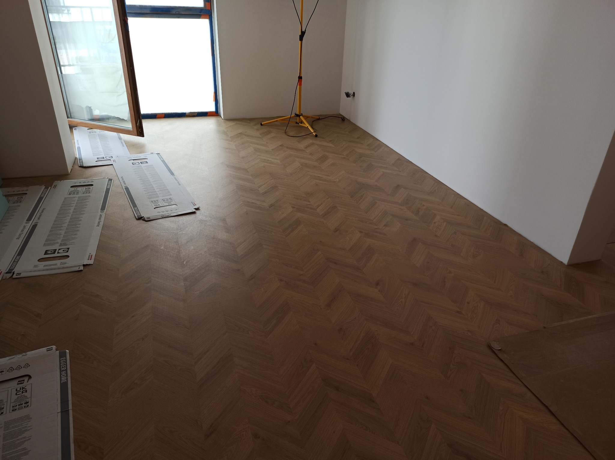 Ułożona, wzorzysta podłoga drewniana w jodełkę, z widocznymi kartonami po panelach i lampą budowlaną w pustym pomieszczeniu z oknem.