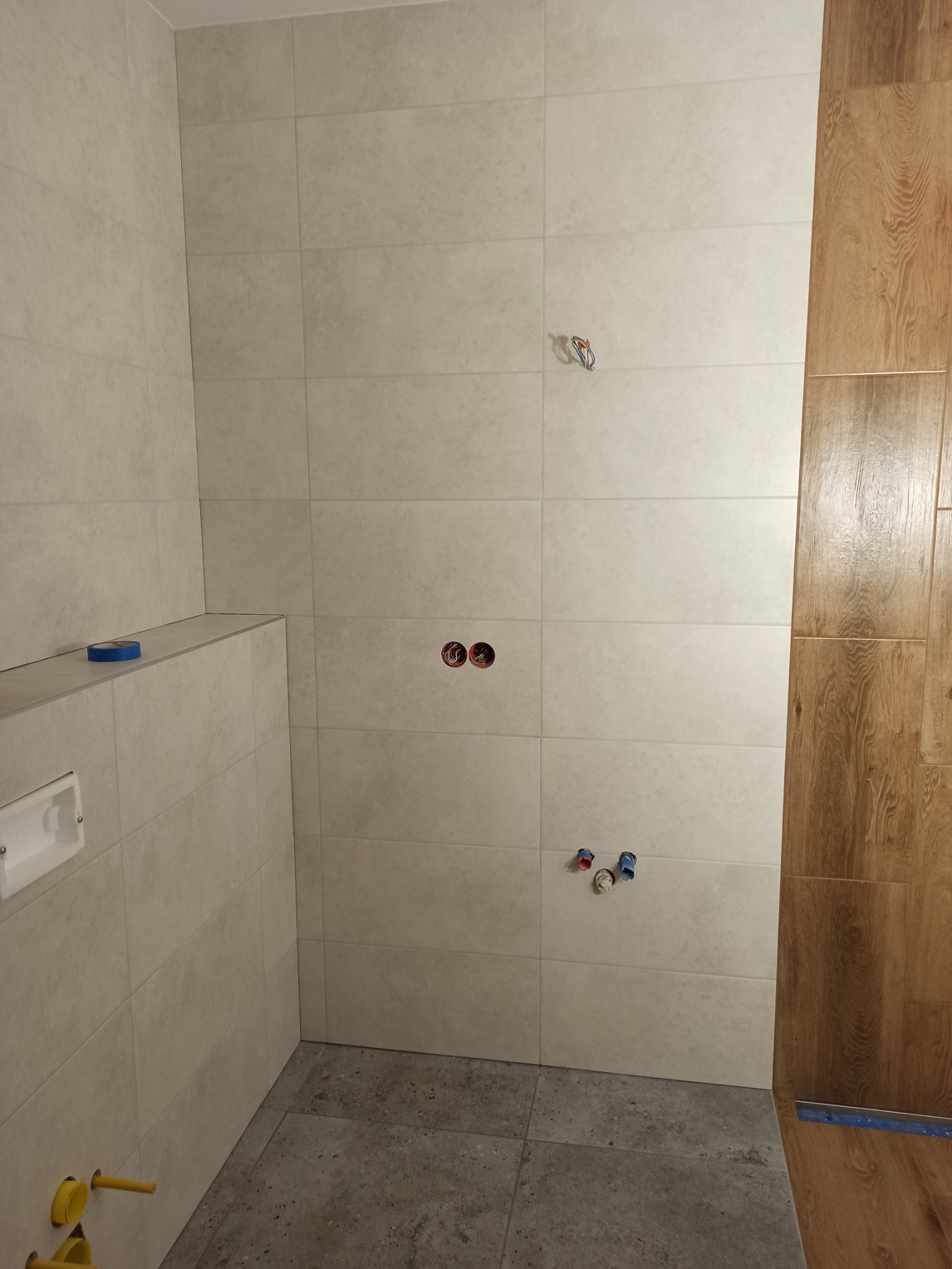 Ściany łazienki wyłożone jasnymi, matowymi płytkami ceramicznymi w trakcie remontu, widoczne przygotowane punkty instalacyjne wodno-kanalizacyjne i elektryczne, fragment drewnopodobnej okładziny...