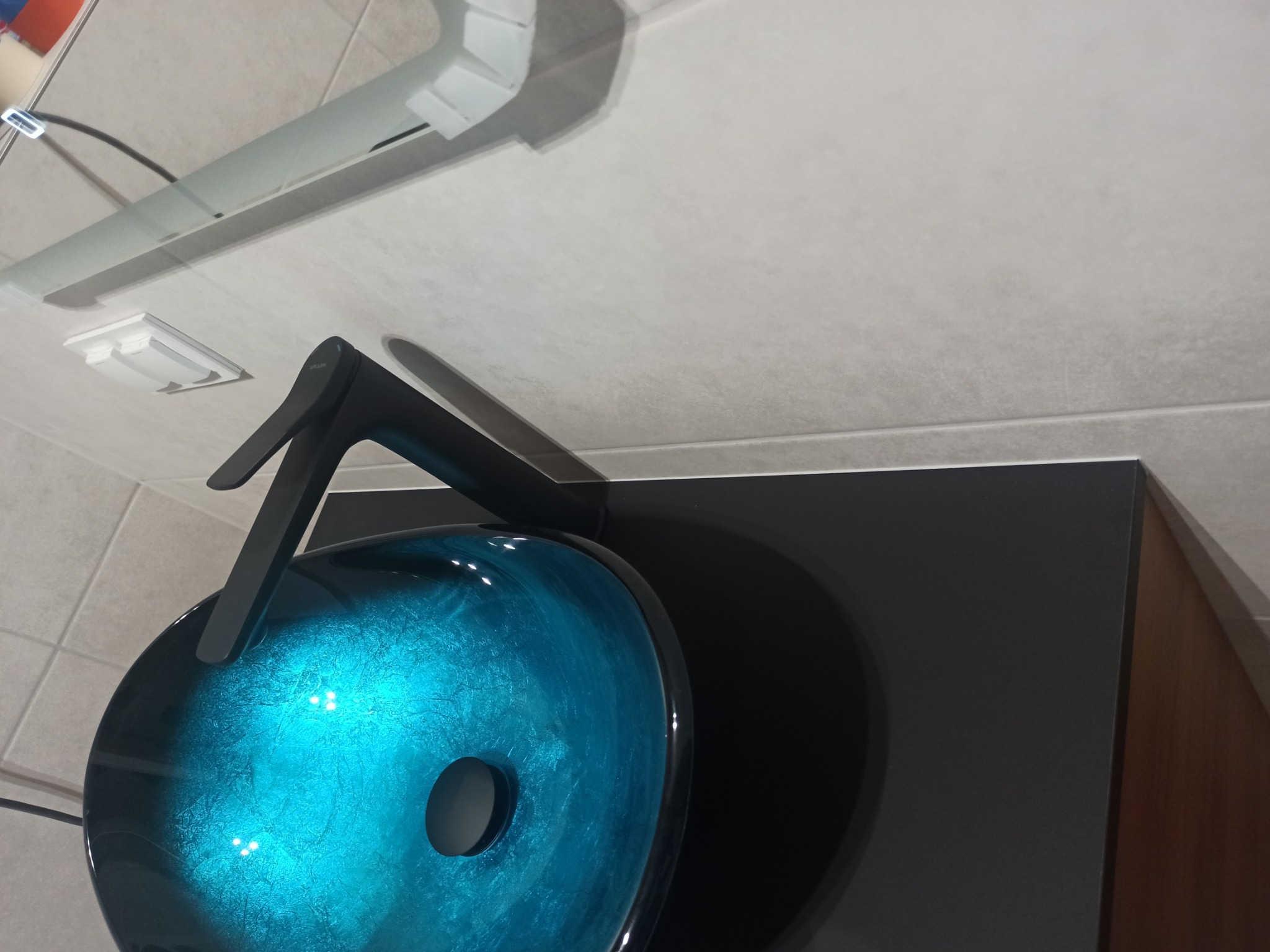 Niebieska, szklana umywalka nablatowa z czarną armaturą na czarnej szafce w łazience z beżowymi płytkami.